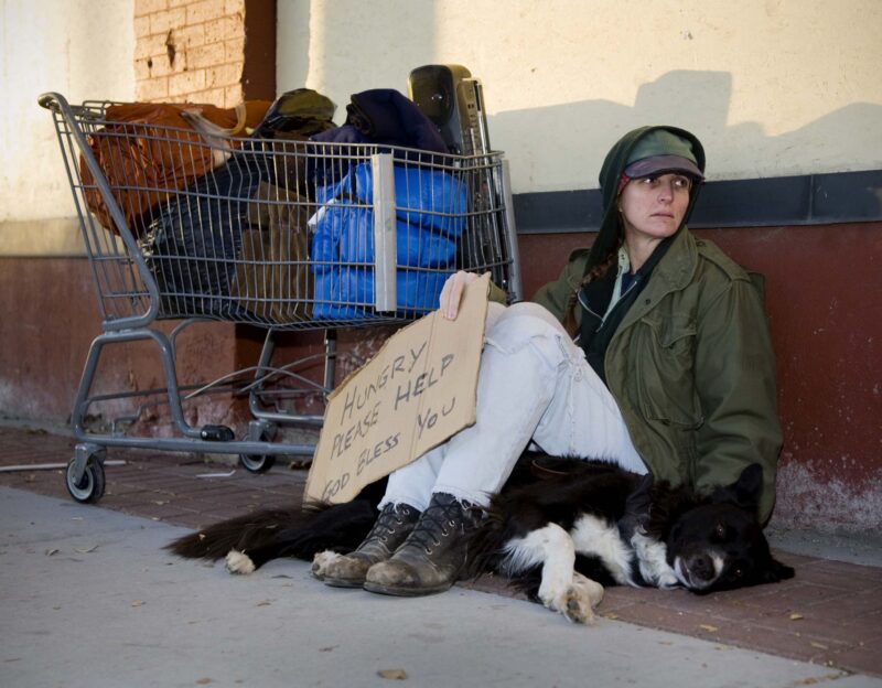 An unhoused woman sitting on a sidewalk with her dog.