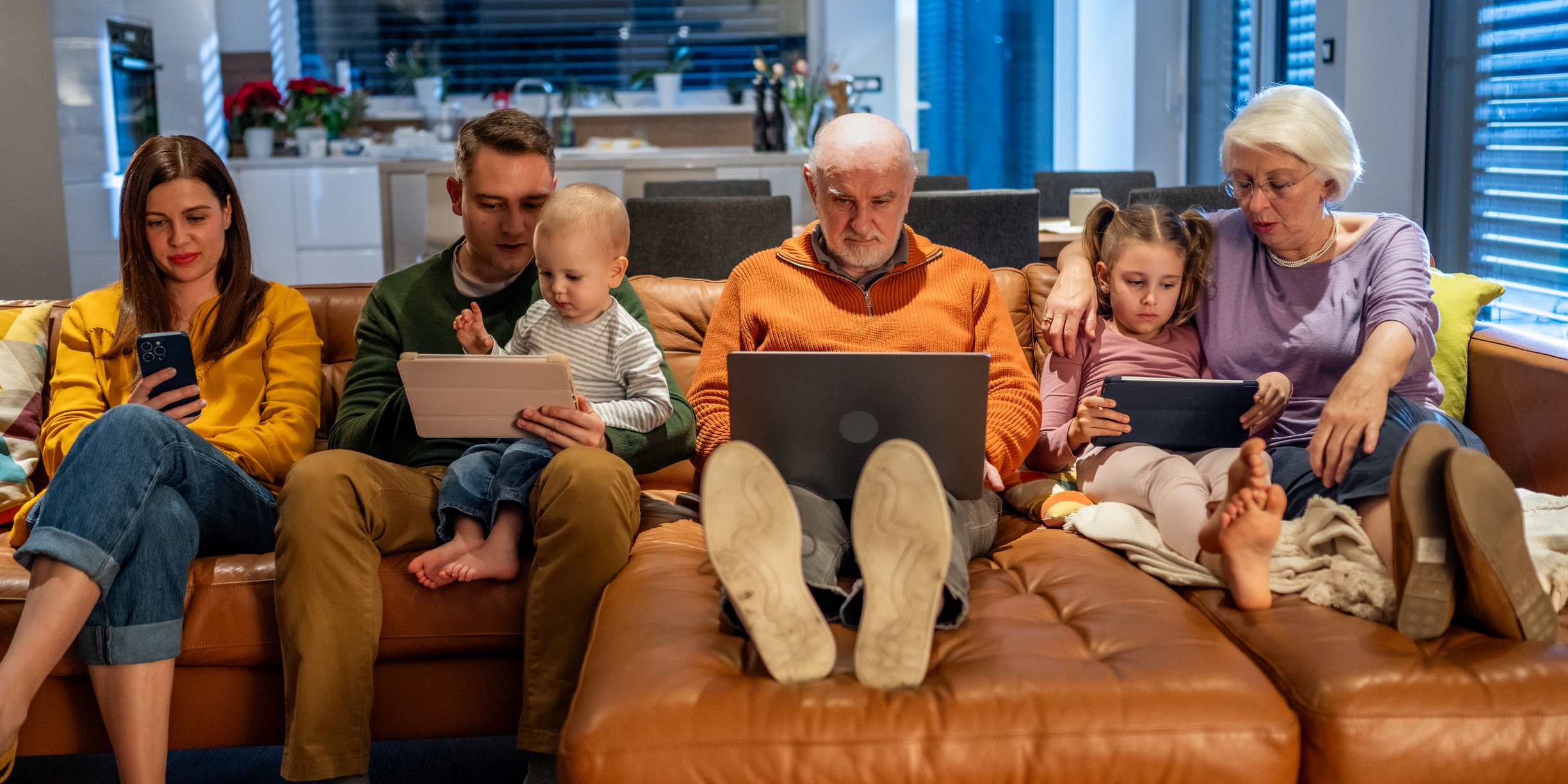 A diverse multigenerational family, including children and seniors, immersed in digital devices while sitting together on a cozy sofa in a warmly lit, modern living room. The background features a tidy, open kitchen area.