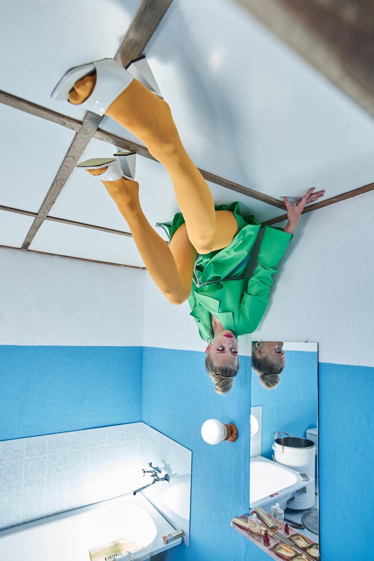 Upside down view of the female model wearing bright clothes sitting at the ceiling and looking away while posing at the strange studio. 