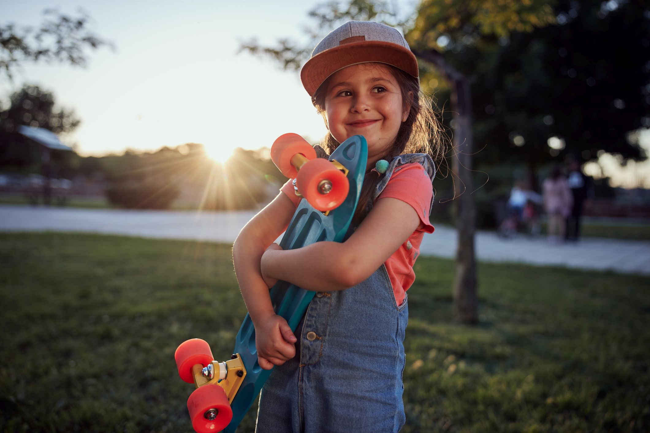Cute little girl holding her small skateboard in a public park during sunset