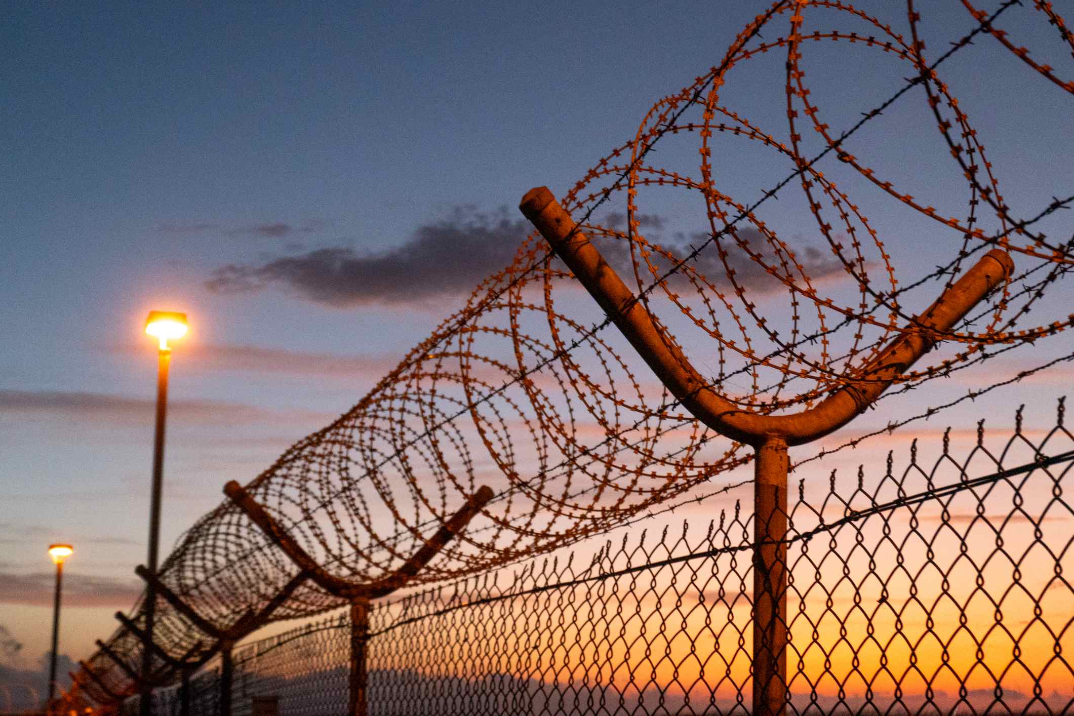 Razor wire, barbed wire, mesh wire fencing and floodlights forming part of the security perimeter of Sydney Kingsford Smith Airport.  This image was taken from near Shep's Mound, Ross Smith Avenue, Mascot on a hot and sunny evening after sunset 