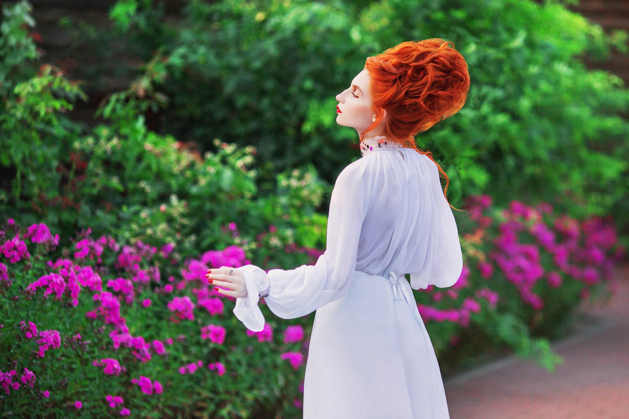 A red-haired woman in an old-fashioned white dress stands in a garden, looking into the distance