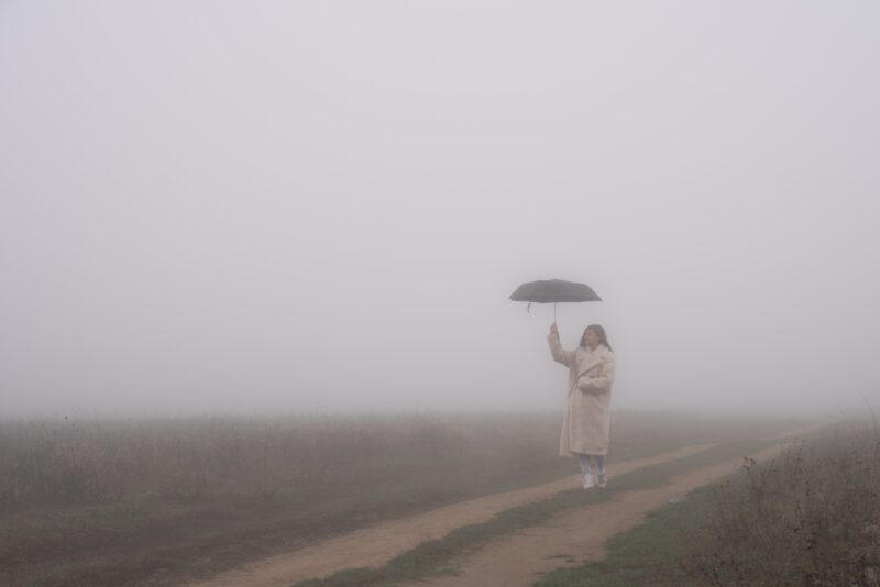 Young girl in wite coat stands in fog among the meadow on a dirt road and holds umbrella in her hand, autumn time