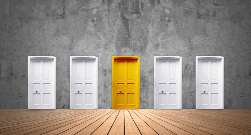 A cement wall with a row of five doors. Four are white, but the one in the middle is yellow.