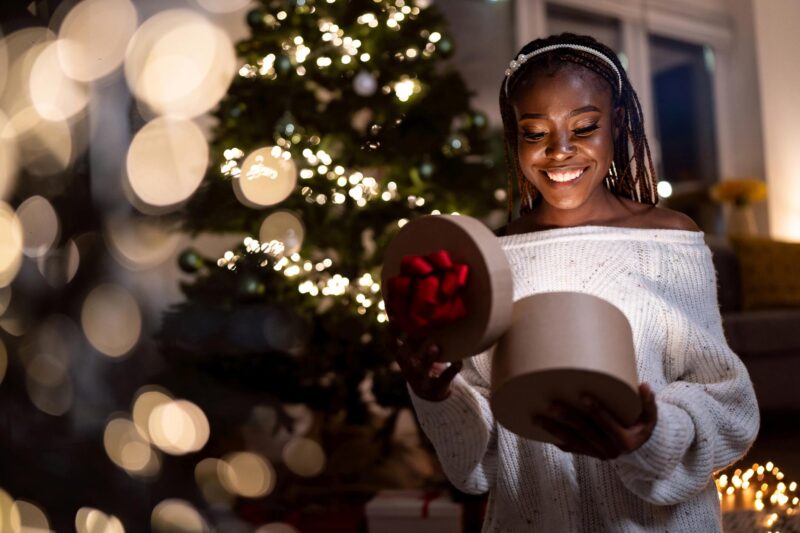 A young black woman opens a present with a lighted tree behind her