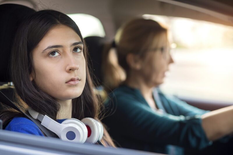 A teen with headphones around her neck rolls her eyes and looks out the window while her mother drives