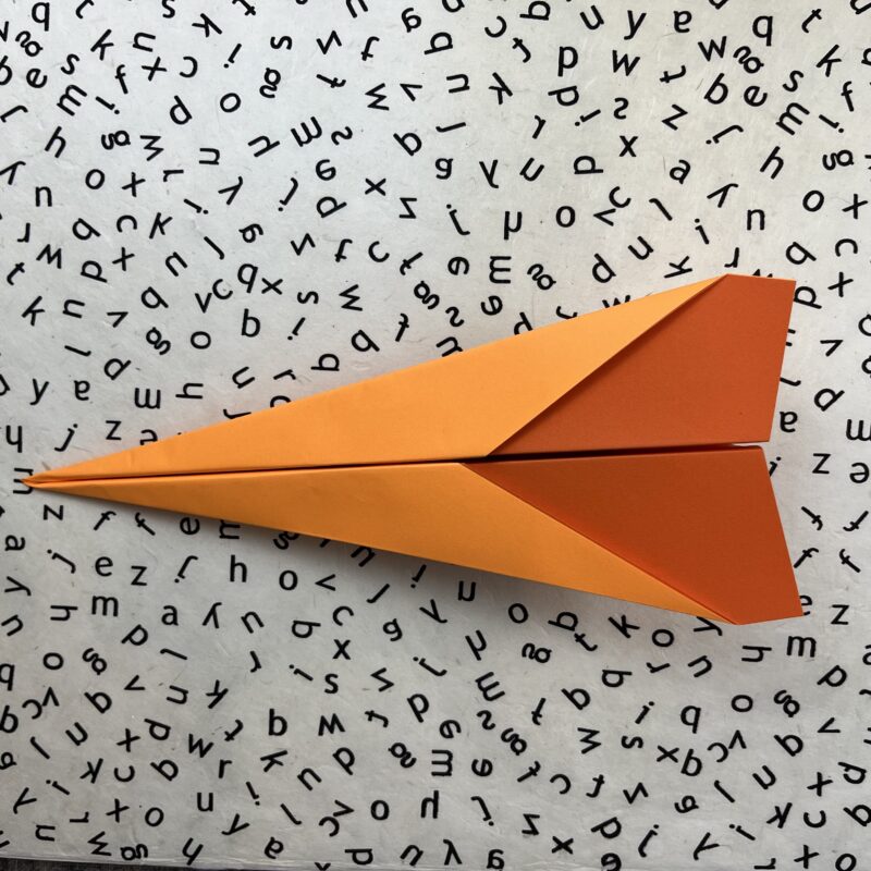 Finished orange dart paper airplane on black and white alphabet background.